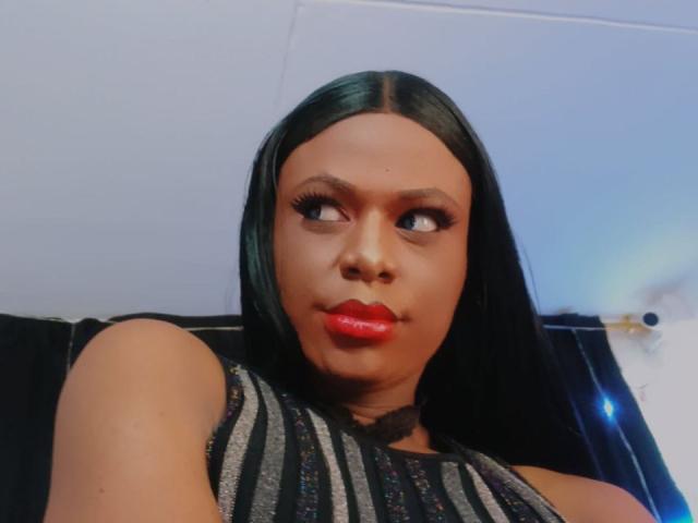 VenusMiller has dark skin, long black hair, red lipstick, and is wearing a sleeveless black and gray striped top, looking to the side with a slightly pouty expression.