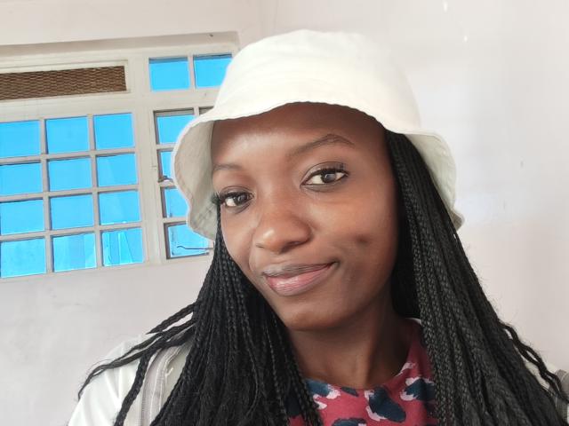 PrettyPice tiene la piel oscura, trenzas negras largas, usa un gorro blanco tipo cubo, y una camisa roja con estampado floral, sonríe ligeramente con lápiz labial rosa, ojos marrones y una linda peca..