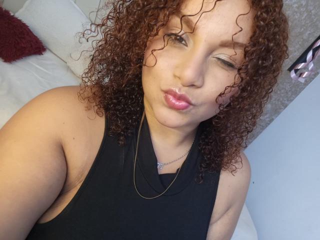 YeraCarey with curly brown hair, light brown skin, wearing a black halter top, making a pouty expression, winking, pink glossy lips, silver necklace, and a subtle smile.
