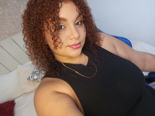 YeraCarey with curly brown hair, black sleeveless top, pink lipstick, and a gold necklace, sitting on a bed, showing her tits and a slight smile.