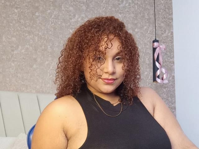 YeraCarey has curly brown hair, light brown skin, and wears a black sleeveless top. She has small tits, a subtle smile, and pink lipstick. Background is grey and white.
