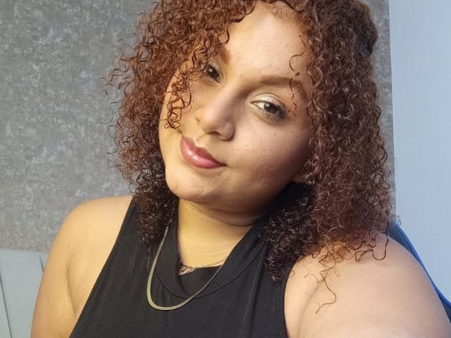 YeraCarey has curly brown hair, light skin, and wears a black sleeveless top with a gold necklace. She's smiling with pink lips and looking slightly to the side.