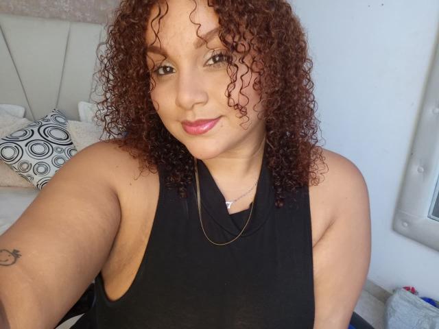 YeraCarey with curly red hair, black halter top, pink lipstick, small tattoo on left arm, necklace, smiling, medium tits, light skin, bedroom, daytime.