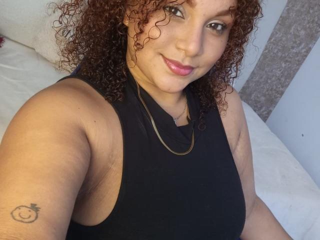 YeraCarey with curly brown hair, light brown skin, pink lipstick, black sleeveless top, gold necklace, smiling, small tattoo of a smiling face on upper left arm.