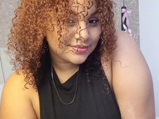 YeraCarey with curly red-brown hair, light brown skin, wearing a black sleeveless top, gold necklace, and pink lipstick, winking with a slight smile, showing her shoulder.