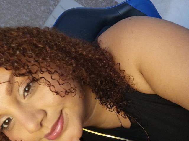 YeraCarey with curly brown hair, smiling, wearing a black top, showing her bare shoulder and ass, lying on a blue and black chair.