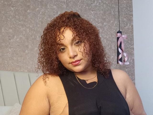 YeraCarey has curly brown hair, light brown skin, and is wearing a black sleeveless top. Her tits are visible through the side cutout. She's smiling slightly.
