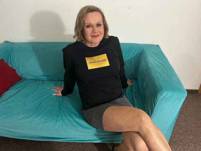 KatharinaSky-hot, blonde, sitting on teal couch, black sweater, gray skirt, crossed legs, smiling, red pillow, white wall, carpeted floor, tan skin, medium tits, relaxed, confident.