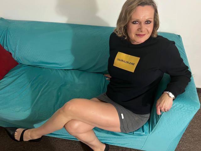 KatharinaSky-hot with medium tits, gray skirt, black sweater, and sandals, sitting on a turquoise couch, legs crossed, smiling at the camera. She's wearing a white wristwatch.