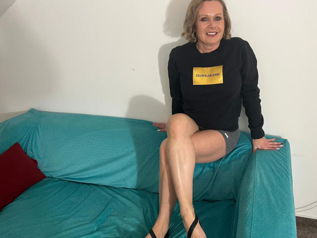 KatharinaSky-hot, middle-aged woman with blonde hair, black sweater, gray shorts, and black heels, sitting on a teal couch with crossed legs, smiling at the camera.