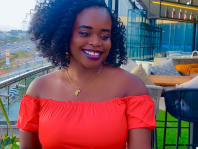 HeadLuna with curly black hair, wearing a red off-shoulder top, showing her boobs, smiling with purple lipstick, gold necklace, and dark skin, sitting on a balcony.
