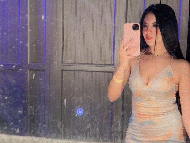 EimmyPark with long black hair, wearing a tight, shiny, pastel dress with a deep V, holding a pink phone, smiling at herself, showing her boobs, in a mirror selfie.