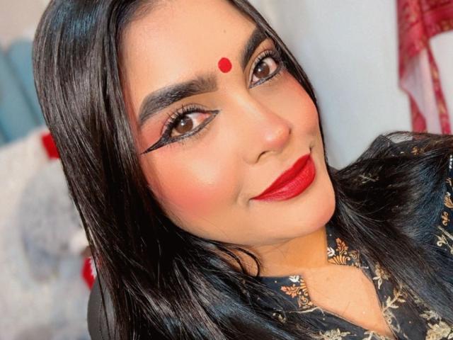 LakshmIrani has dark black hair, brown eyes, red lipstick, and a red dot on her forehead. She's wearing bold black eyeliner and a dark floral-patterned shirt.