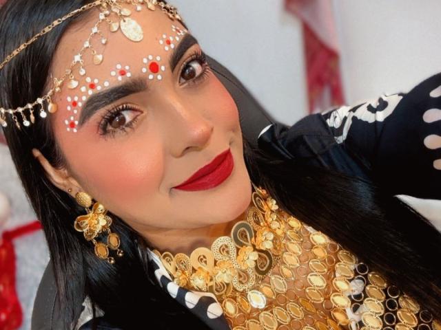 LakshmIrani has dark hair, red lipstick, and a gold headpiece with red and white gems. She's wearing a black, white, and gold outfit with a massive gold necklace.