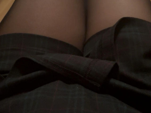 SugaryCarrie's dark plaid skirt is bunched up, revealing her black pantyhose-covered thighs. The skirt's fabric has a subtle check pattern, creating a slight texture.