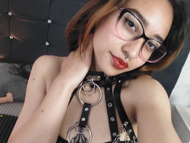 CamiVargas with glasses, nose ring, and red lipstick, wearing a black leather harness with metal rings, touching her neck, short brown hair with orange highlights.