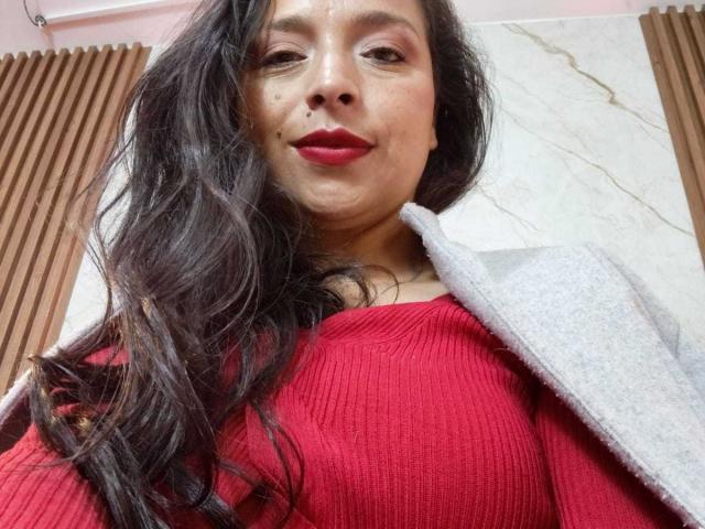StangelarRivera with long black hair, red lipstick, and red ribbed top, wearing a gray coat, smiling slightly, showing small moles on her face.