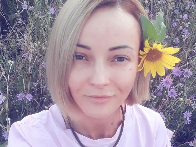 SophieCrach has blonde bob, light skin, and a small beauty mark on her right cheek. She's wearing a white shirt with a yellow sunflower in her hair.