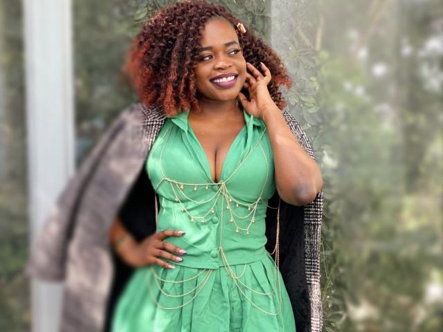 HeadLuna with curly red hair, green dress, gold chain detail, smiling, hand on cheek, black and gray jacket, standing, visible breasts, green eyes, purple lipstick.