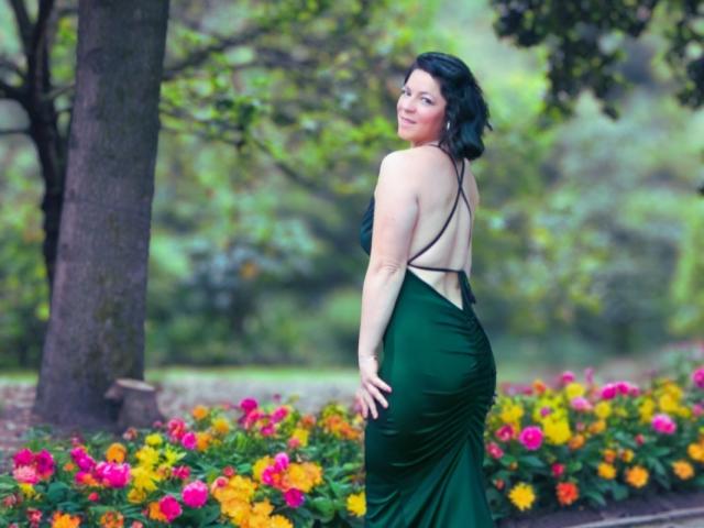 Daisy, gekleed in een groene, rugloze jurk, waarbij haar achterwerk zichtbaar is, staat naast kleurrijke bloemen, kijkt over haar schouder met haar zwarte, golvende haar, in een weelderige, natuurlijke omgeving..