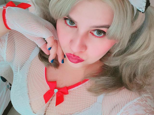 Akkanee with pale skin, blonde hair in pigtails, wearing white mesh top with red ribbon, fishnet gloves, blue nails, red eyeliner, and red lipstick, looking up. Boobs visible.