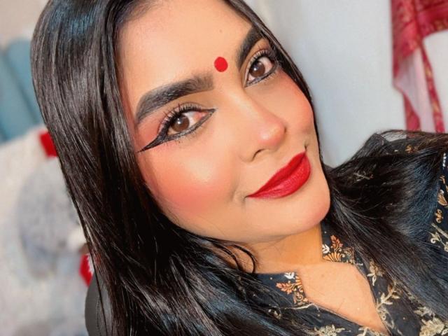 LakshmIrani has dark, shiny hair, red dot on forehead, bold eyeliner, red lipstick, and is wearing a black, floral-patterned shirt. Her ass is not visible.