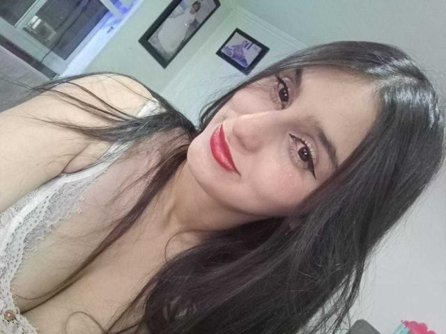 ArheneaLover has long black hair, red lipstick, and is smiling with brown eyes. She's wearing a white lace top, showing her cleavage. The photo is a selfie.