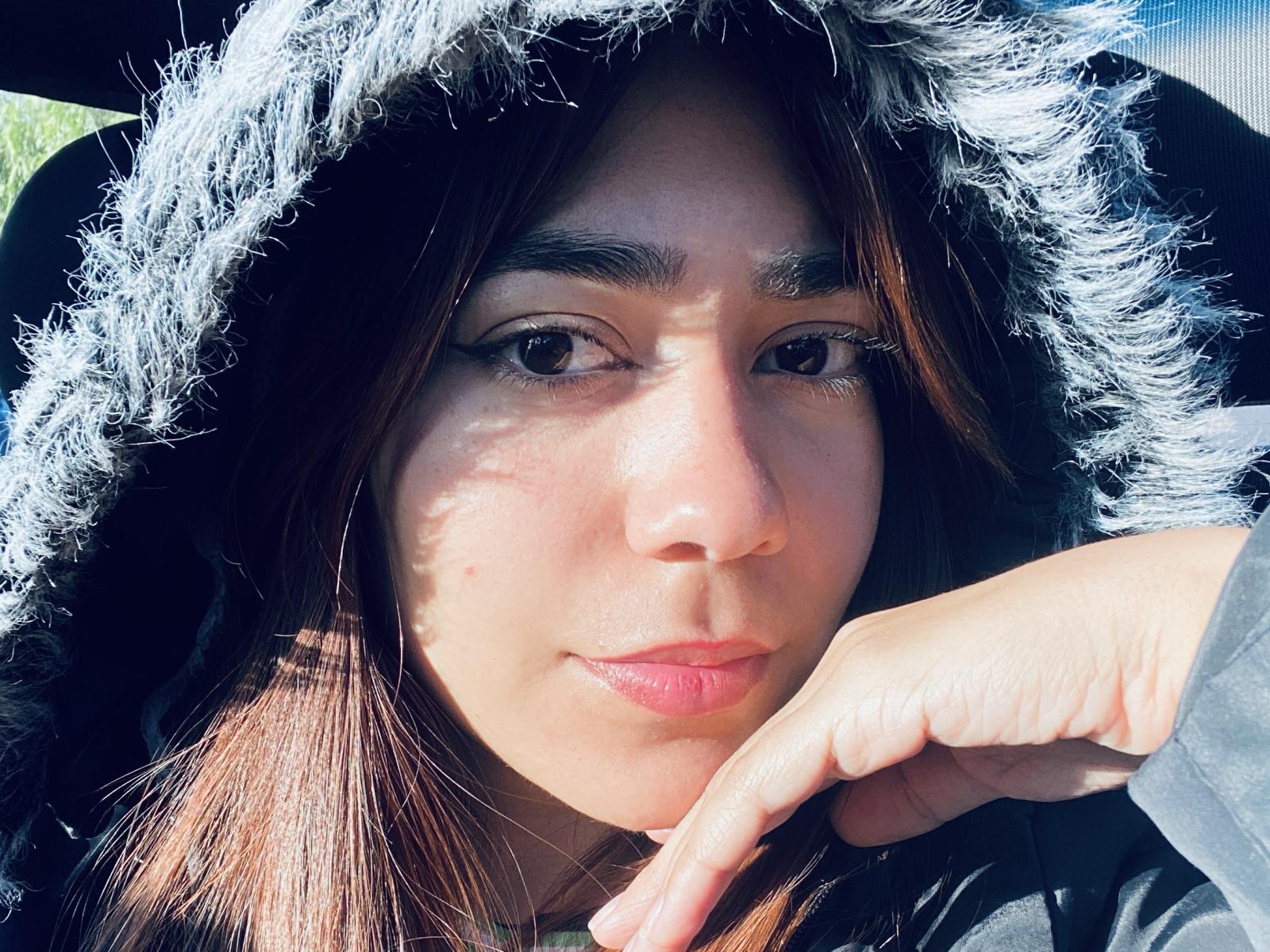 BiaaCam has a cute face with brown eyes, wearing a fluffy black hood, and a subtle smile, with her finger touching her chin in a sunlit selfie.