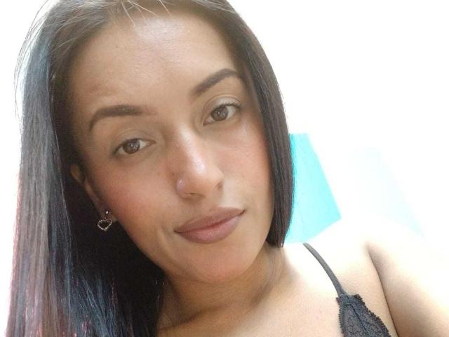NicolleGlory has medium brown skin, black straight hair, brown eyes, small heart earrings, black lace top, and a subtle smile showing her small, natural tits.
