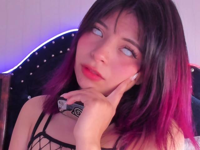 ShofiaWett with pink and black hair, blue eyes, and red lips, holding a camera, wearing a black fishnet top, touching her cheek, looking slightly away.