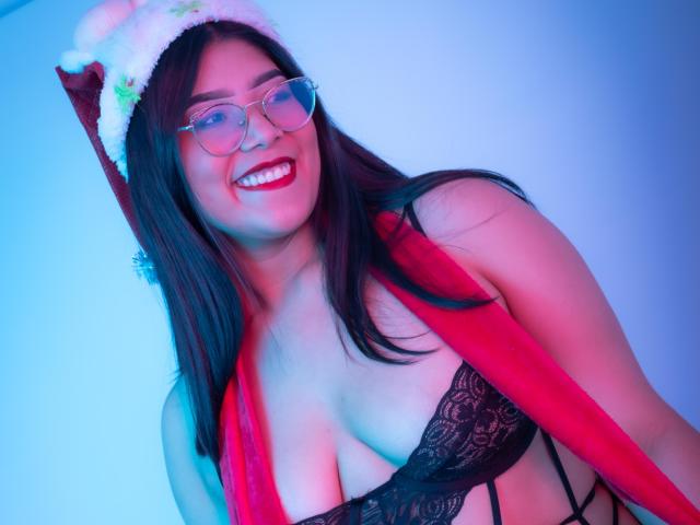 AlexaKiim with red Santa hat, glasses, and red scarf, smiling, showing black lace bra, exposing her big tits, and long brown hair, in blue and pink lighting.