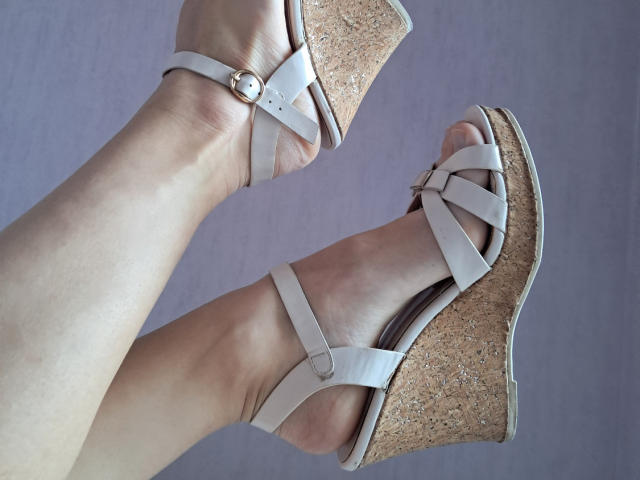 Amberia's pale legs in beige wedge sandals with straps and a small buckle, showing her toes and the cork heel, against a plain background.