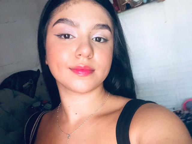 SammyMorra with long black hair, pink eyeshadow, red lipstick, and a silver necklace, wearing a black top, smiling slightly. Her tits are small, and she has a cute face.