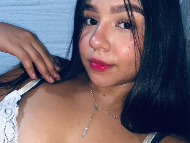 SammyMorra with shiny brown skin, long black hair, pink lipstick, white lace top, necklace with cross, lying down, hand near face, soft smile, slightly flushed cheeks, sexy.