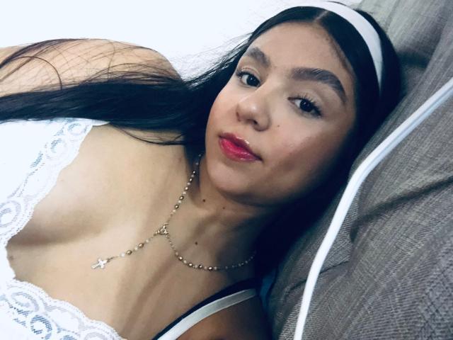 SammyMorra with long black hair, white lace dress, red lipstick, white headband, necklace, lying on gray pillow, showing small tits and a cute face.