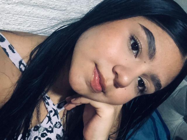 SammyMorra with black hair, brown eyebrows, and shiny pink lips, resting her head on her hand, wearing a white floral top, looking at the camera.