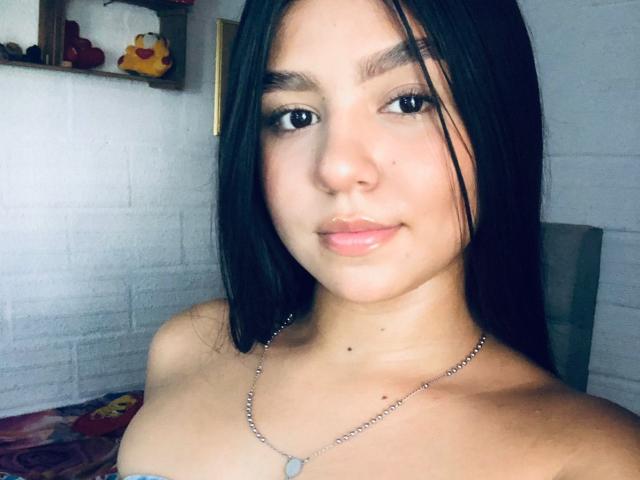 SammyMorra with long black hair, brown eyes, and a small nose, wearing a silver necklace with a small pendant, showing bare shoulders and a subtle smile.