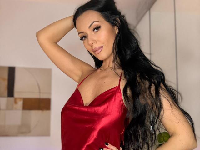 Nadeen with long black hair, red satin dress, and hand in hair, showing off her tits and a playful tongue-out expression. Her makeup is glamorous, and she's sexy.