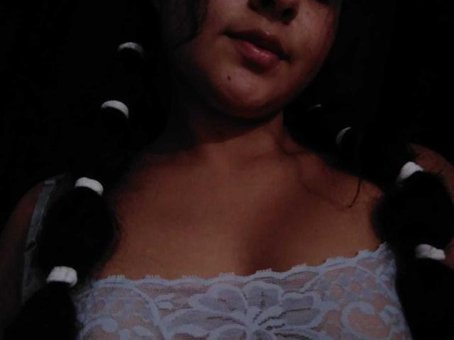 ScarlettZ has dark braids with white bands, wearing a sheer white lace top that shows her small tits. Her brown skin is dimly lit, with a small beauty mark.