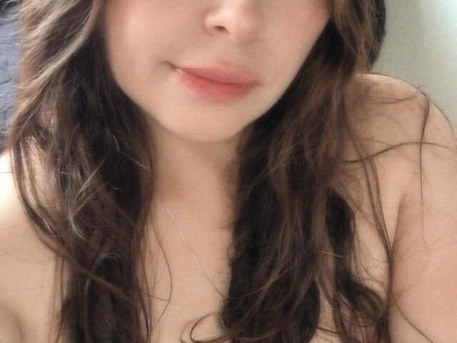 Seher with wet brown hair, pale skin, and pink lips, showing a slight smile, with a silver necklace, and no clothes visible, focusing on her upper chest and face.