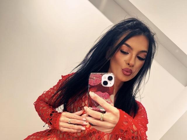 KinkyAmelie with long black hair, red lace dress, and red lipstick, taking a mirror selfie with her phone, showing off her ringed fingers and subtle makeup.