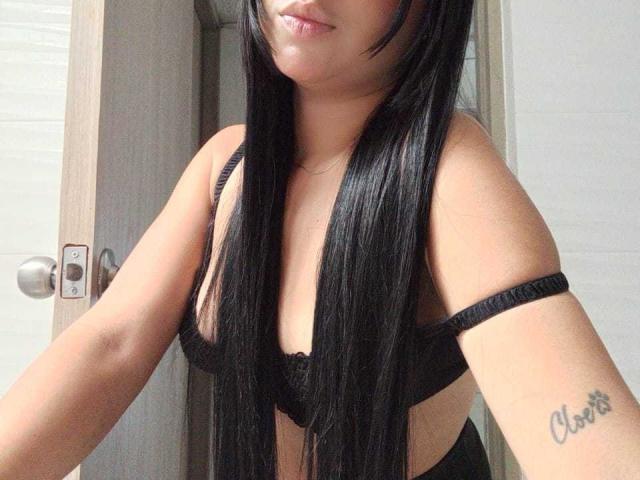 AliceSmity with long black hair, wearing a black bra, showing a tattoo on her right arm that says "Chaz." Her tits are partially visible, and she's smiling slightly.