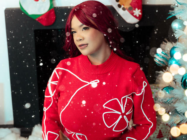VictoriaField with red hair, wearing a red sweater with white floral patterns, standing next to a decorated Christmas tree, snowing, looking to the side, tits visible under the sweater.