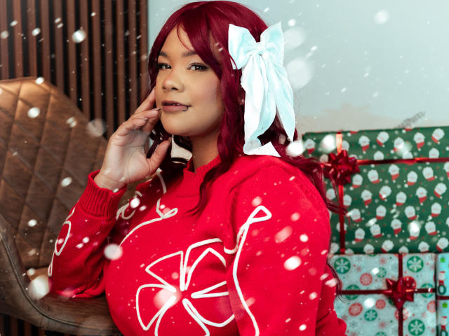 VictoriaField has red hair with a white bow, wearing a red sweater with white Christmas tree patterns. She's looking to the side with a slight smile, her right hand touching her face. Snow-like white dots are scattered across the image. She's seated on a brown quilted chair. The background has green and red patterned gift boxes. The image has a watermark in the upper right corner. The composition emphasizes VictoriaField's red and white outfit against the festive background.