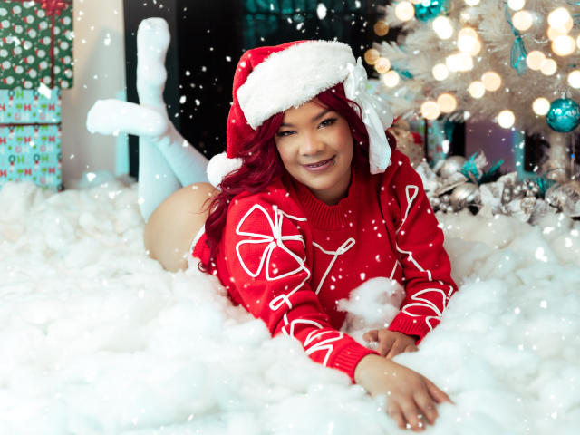 VictoriaField, a woman with red hair, lies on a fluffy white surface, wearing a red Santa sweater with white knit patterns and a Santa hat. She has a cute smile and is positioned with her legs bent and feet up, showcasing white knee-high socks. A white Christmas tree with blue and silver ornaments is in the background. Snowflakes are falling. The image has a festive, holiday feel.