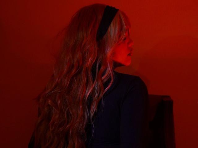 TessaMagic with long wavy blonde hair, black headband, and dark top, illuminated by red light, facing sideways, showing her profile and slightly turned ass.