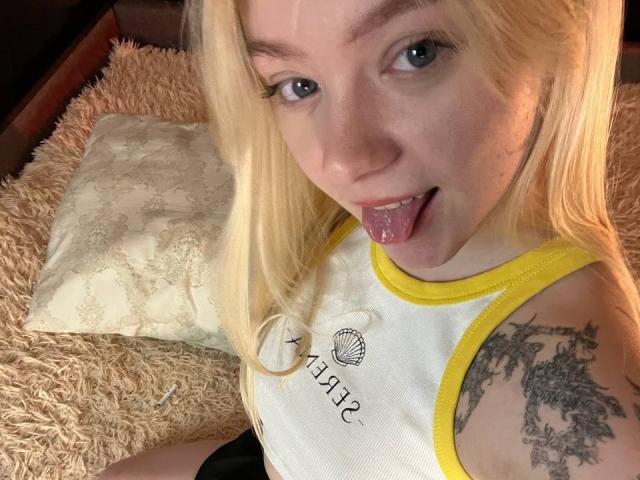 EnchantingEyes with blonde hair, blue eyes, and tongue out, wearing a yellow-trimmed white tank top, showing a tattoo on her right arm, sitting on a fluffy beige pillow.