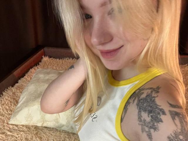 EnchantingEyes with blonde hair, smiling, wearing a yellow-trimmed white tank top, showing a tattoo on her right arm, lying on a fluffy beige pillow.