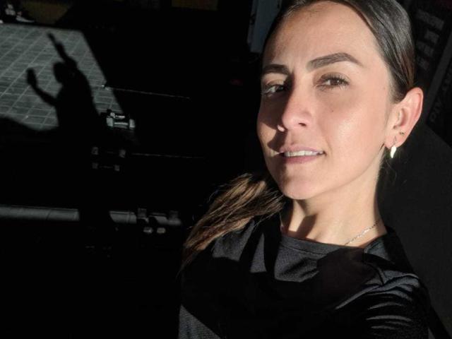 AriaRous with light skin, brown eyes, and a slight smile, wearing a black shirt, has her brown hair in a ponytail, and a silver earring, shadowed on the right.