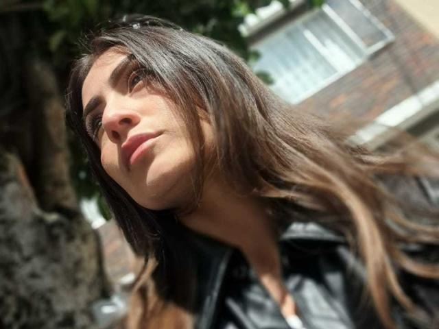 AriaRous has long brown hair, a slight smile, and is looking up with a relaxed expression. She's wearing a black leather jacket, and the sun is shining.